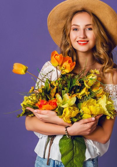 Studio  fashion portrait of pretty cute blonde woman in straw hat, white cotton  shirt sitting and holding bouquet of amazing spring  flowers. Wearing stylish retro  outfit .