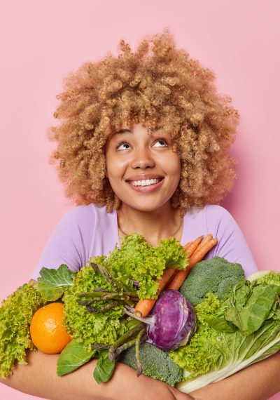 Vertical shot of happy curious woman smiles toothily looks above notices something appealing embraces big bunch of fresh green vegetables keeps to healthy diet isolated over pink background.